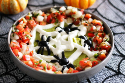Enjoy spooky snacking with this Halloween Spiderweb Avocado Hummus Dip at your Halloween party! This healthy Halloween recipe is made like traditional hummus, but with avocado, a plain Greek yogurt spiderweb, black olive spiders, and pico de gallo!